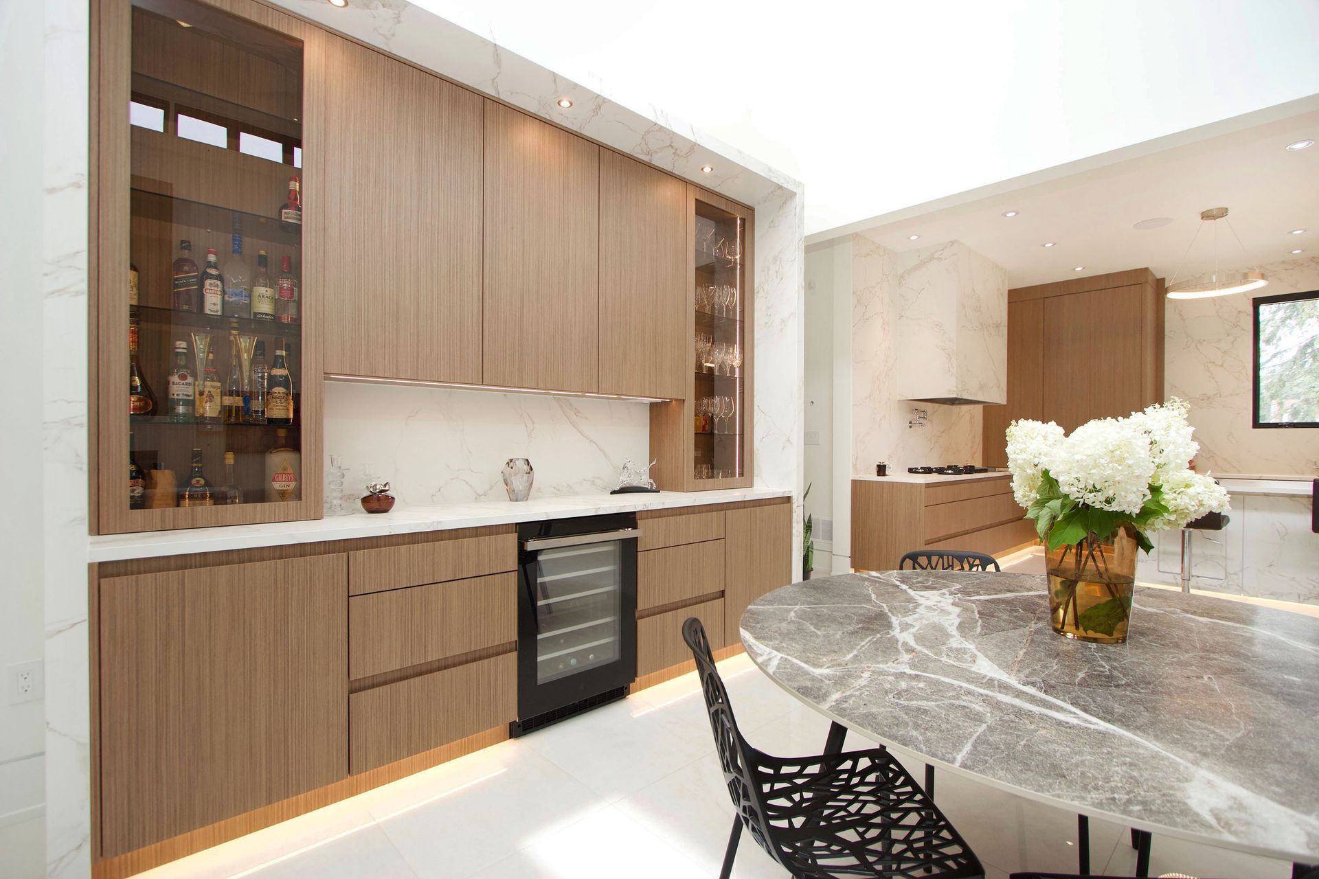 Modern kitchen with wooden cabinetry, marble countertops, and a wine fridge. Round marble table with flowers.
