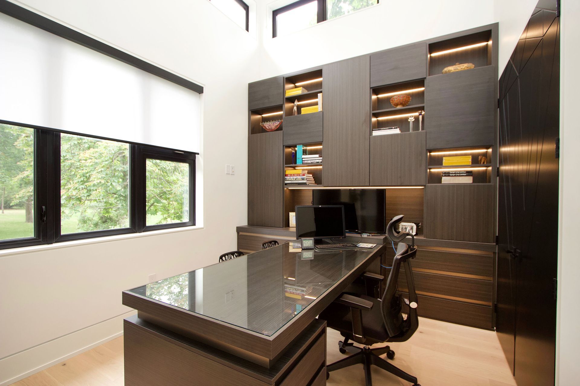 Modern home office with dark wood built-ins, large desk, and rolling chair, overlooking a window.