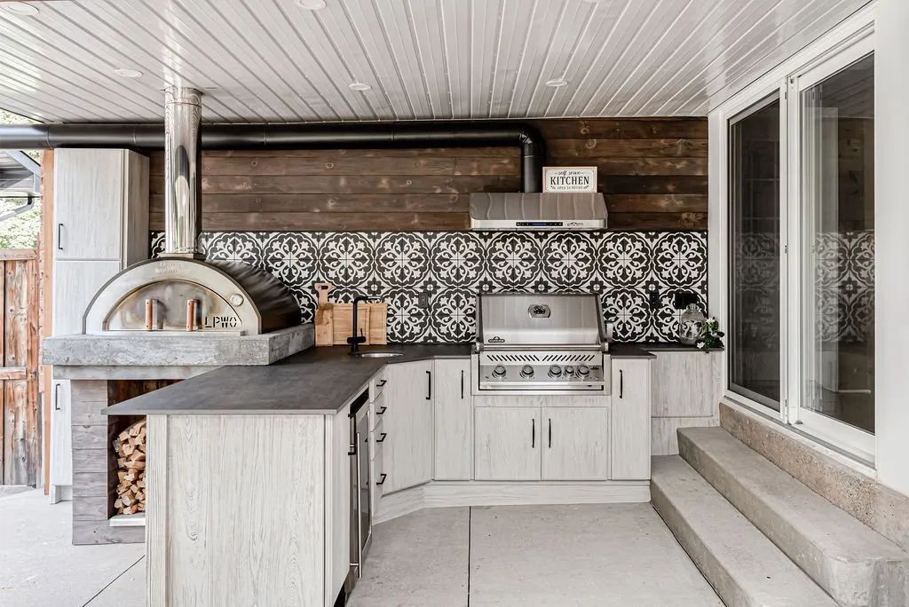 Outdoor kitchen with pizza oven, grill, cabinets, tile backsplash, and wood accent wall.