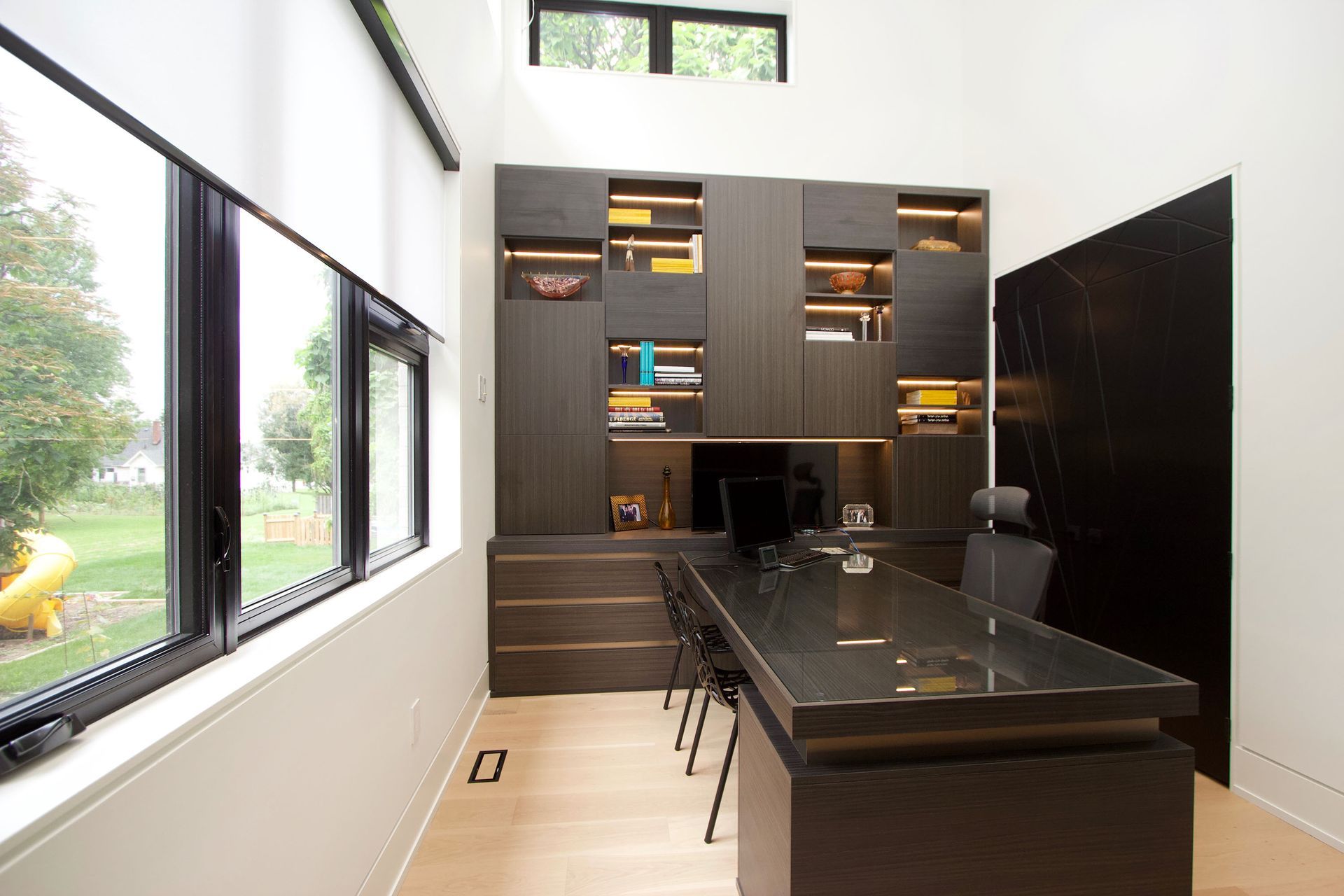 Modern home office: dark wood desk, built-in shelves with lighting, large windows, view of yard.
