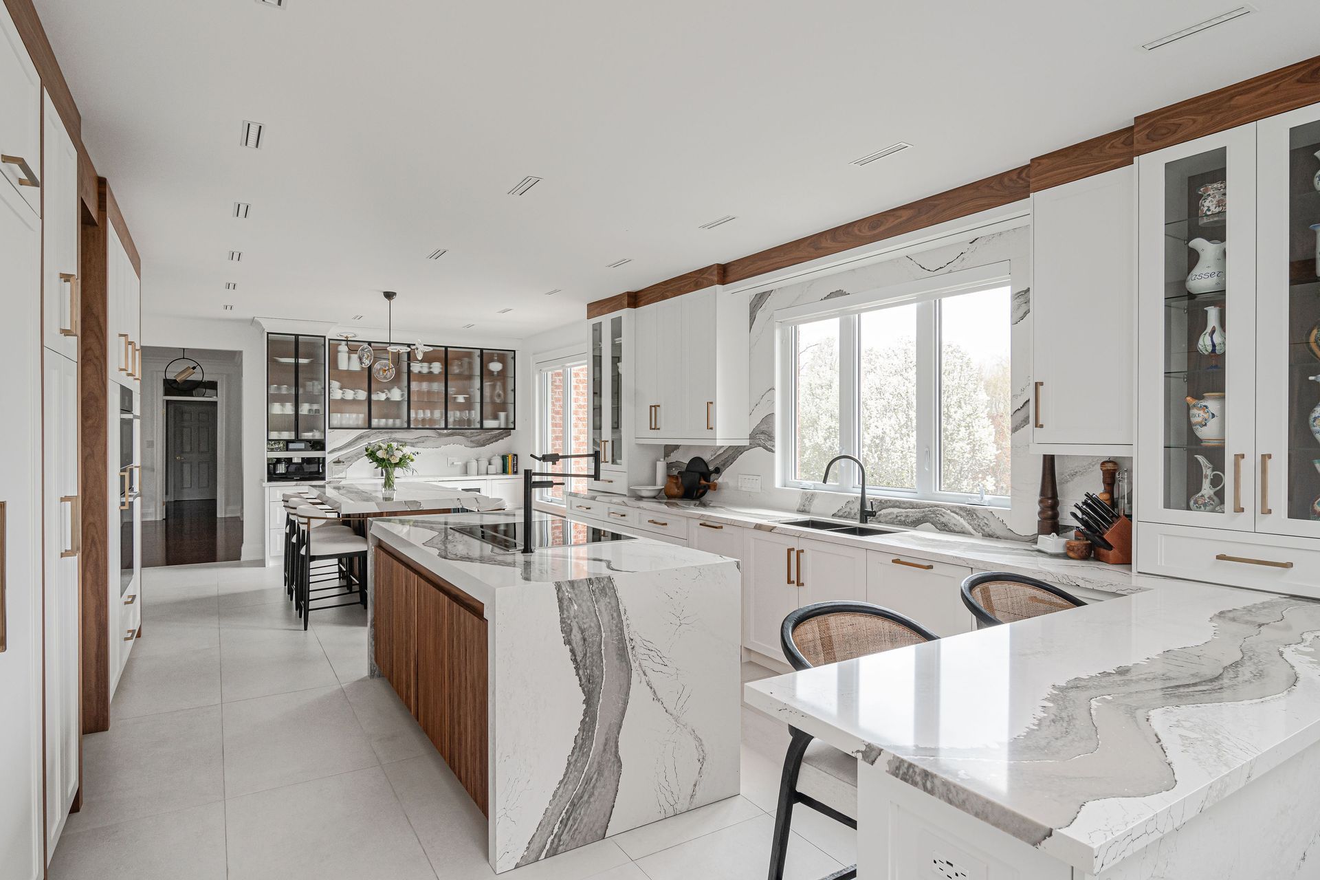 Spacious white kitchen with marble countertops, wooden accents, and island seating; natural light through a large window.