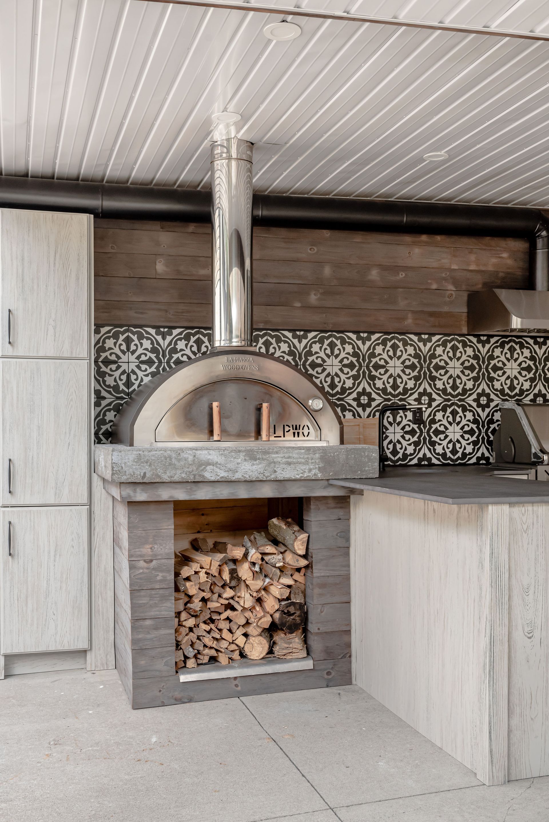 A pizza oven is sitting in the middle of a kitchen next to a pile of wood.
