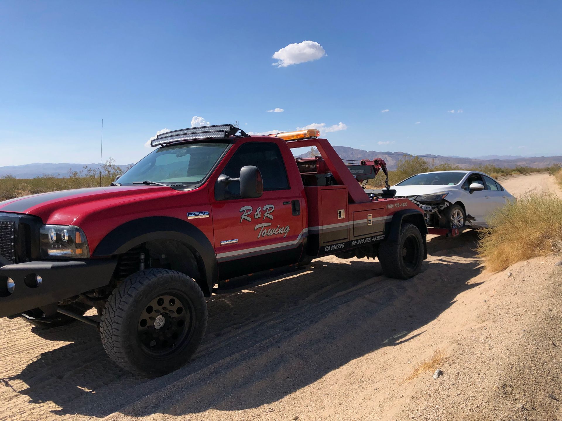 A red tow truck is towing a white car on a dirt road.