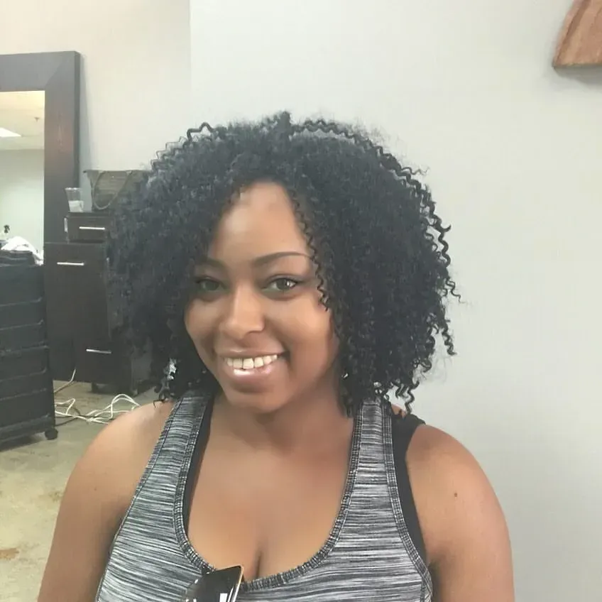 Smiling person with curly hair in a gray sleeveless top, seated indoors.