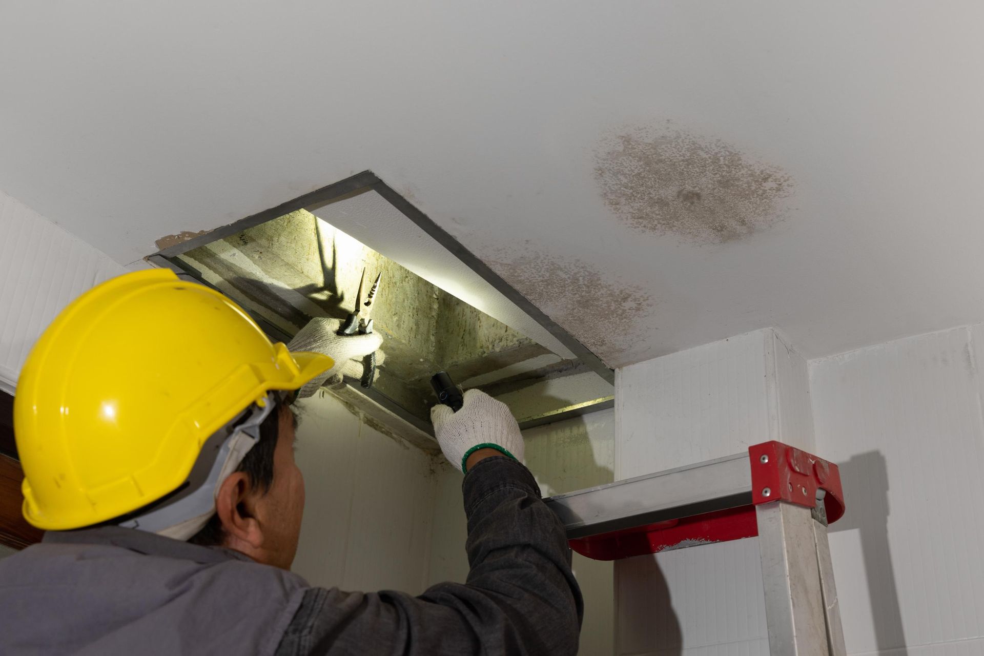 A man wearing a hard hat and gloves is looking through a hole in the ceiling.