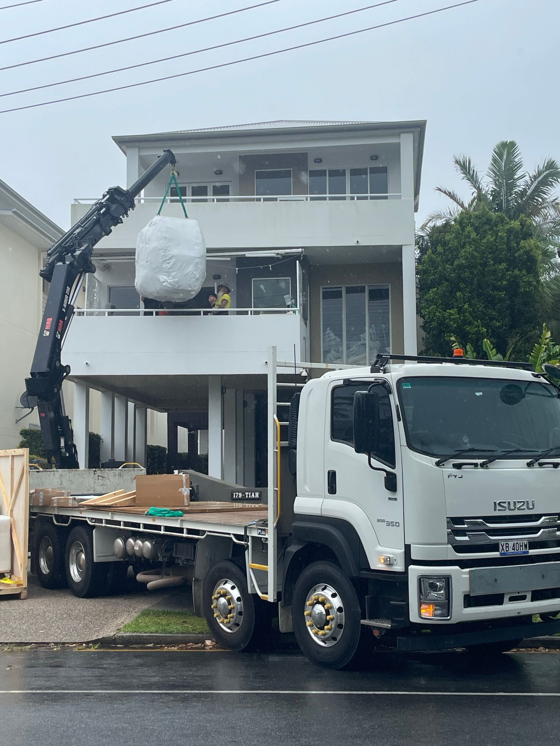 White flatbed crane truck lifting a wrapped item in front of a two-story house during — Trucking Crane Trucks in Warana, QLD