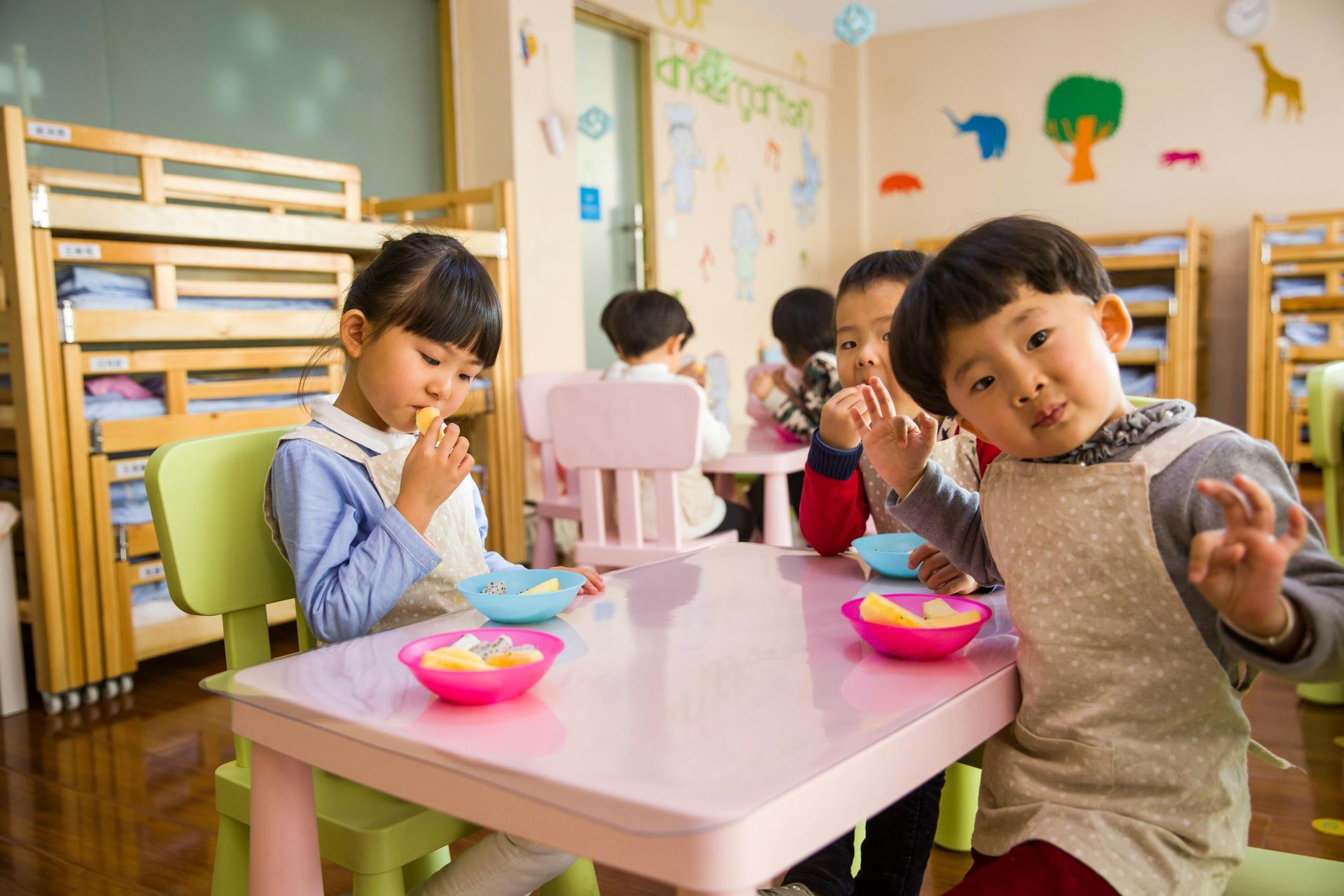 Children eating fruit at a pink table in a colorful classroom. One child gives an “OK” gesture.