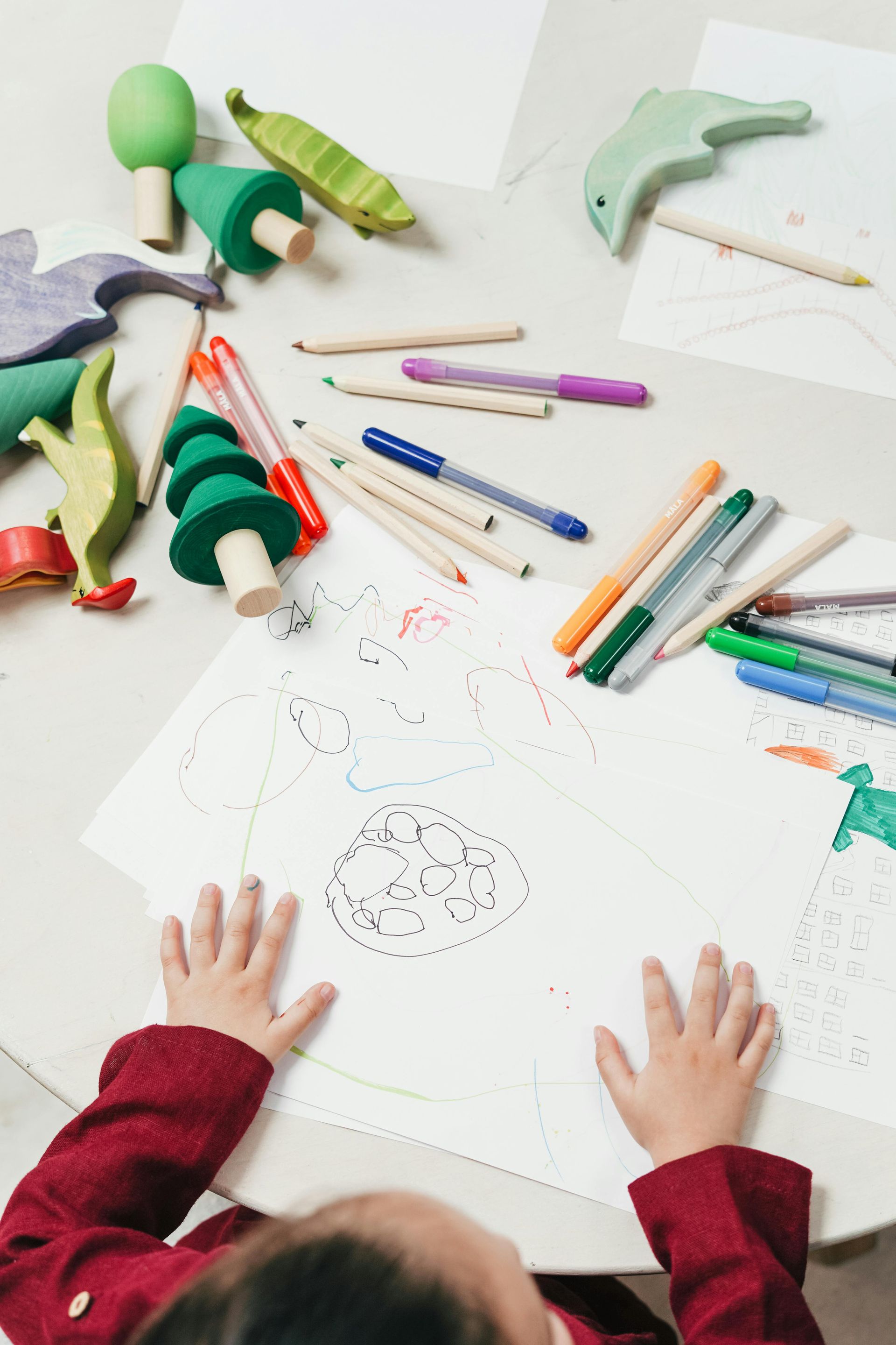 Child drawing at a table with crayons and wooden toys, including trees and a dolphin.