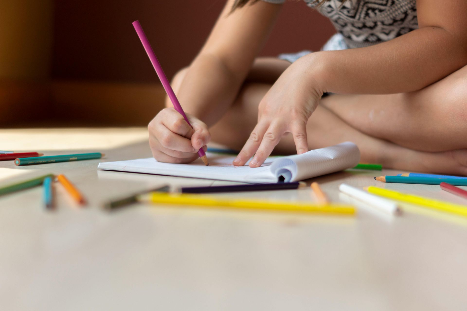 Child drawing with a pink pencil in a notebook, surrounded by colored pencils, sitting on a floor.
