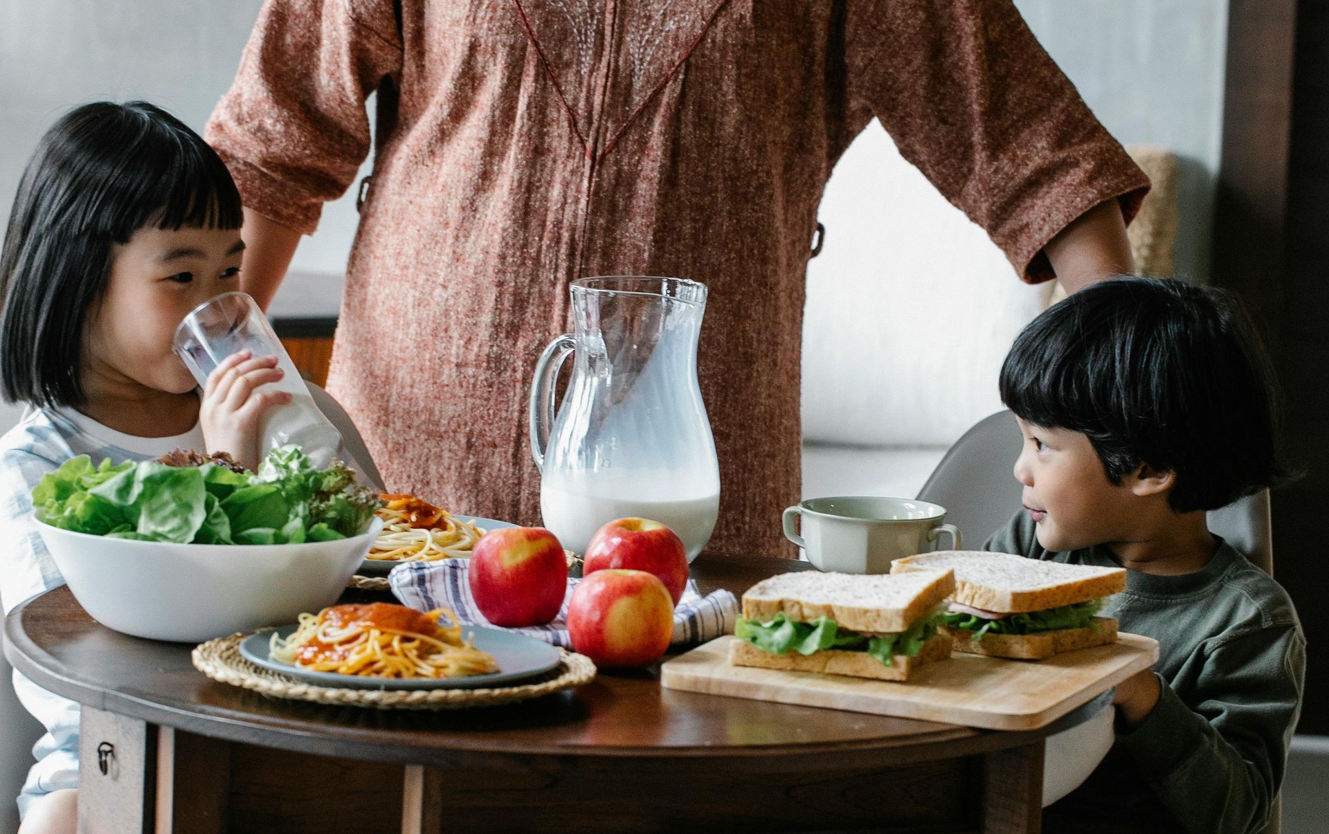 Two children and an adult at a table with food and milk. One child drinks from a glass.