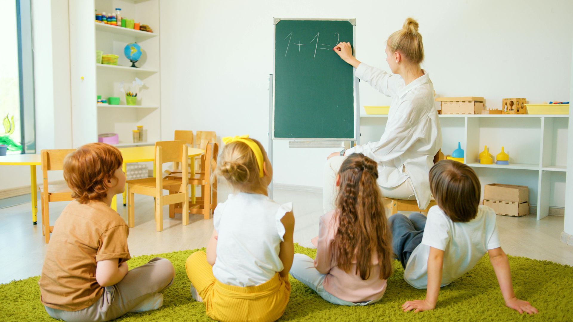 Teacher writing on a chalkboard for a group of children seated on a green rug in a brightly lit classroom.