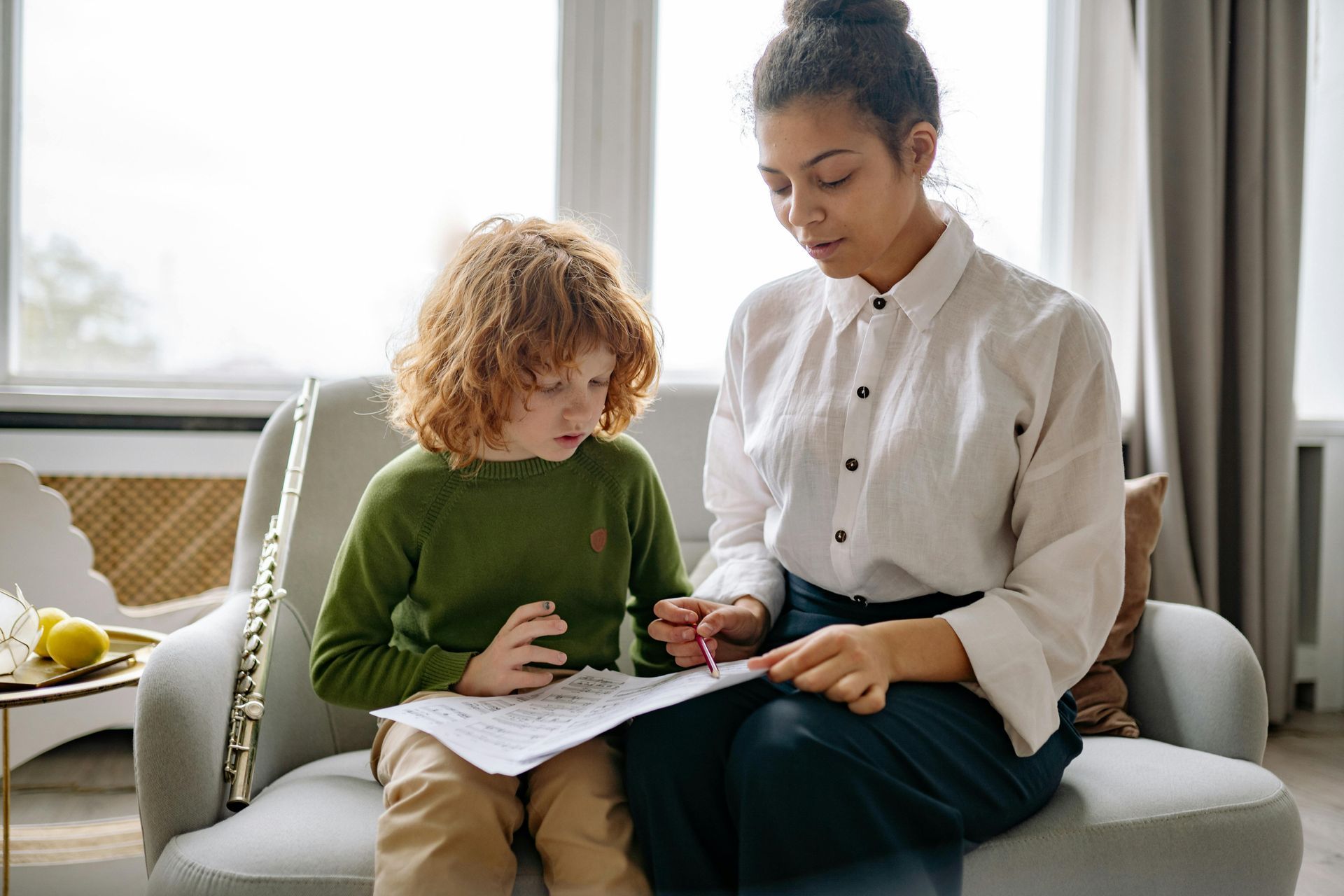 Woman teaching a child, seated on a couch, pointing at sheet music. Light, bright room.