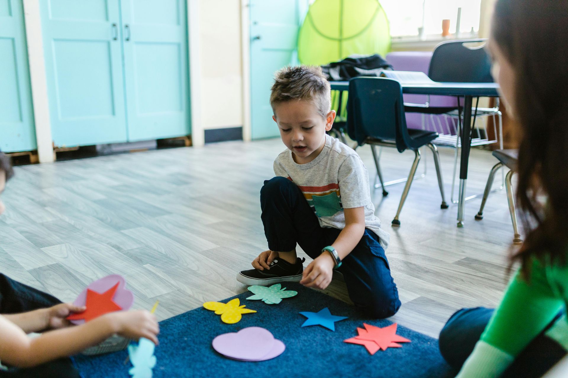 Boy on the floor arranging colorful paper cutouts with an adult in a classroom setting.