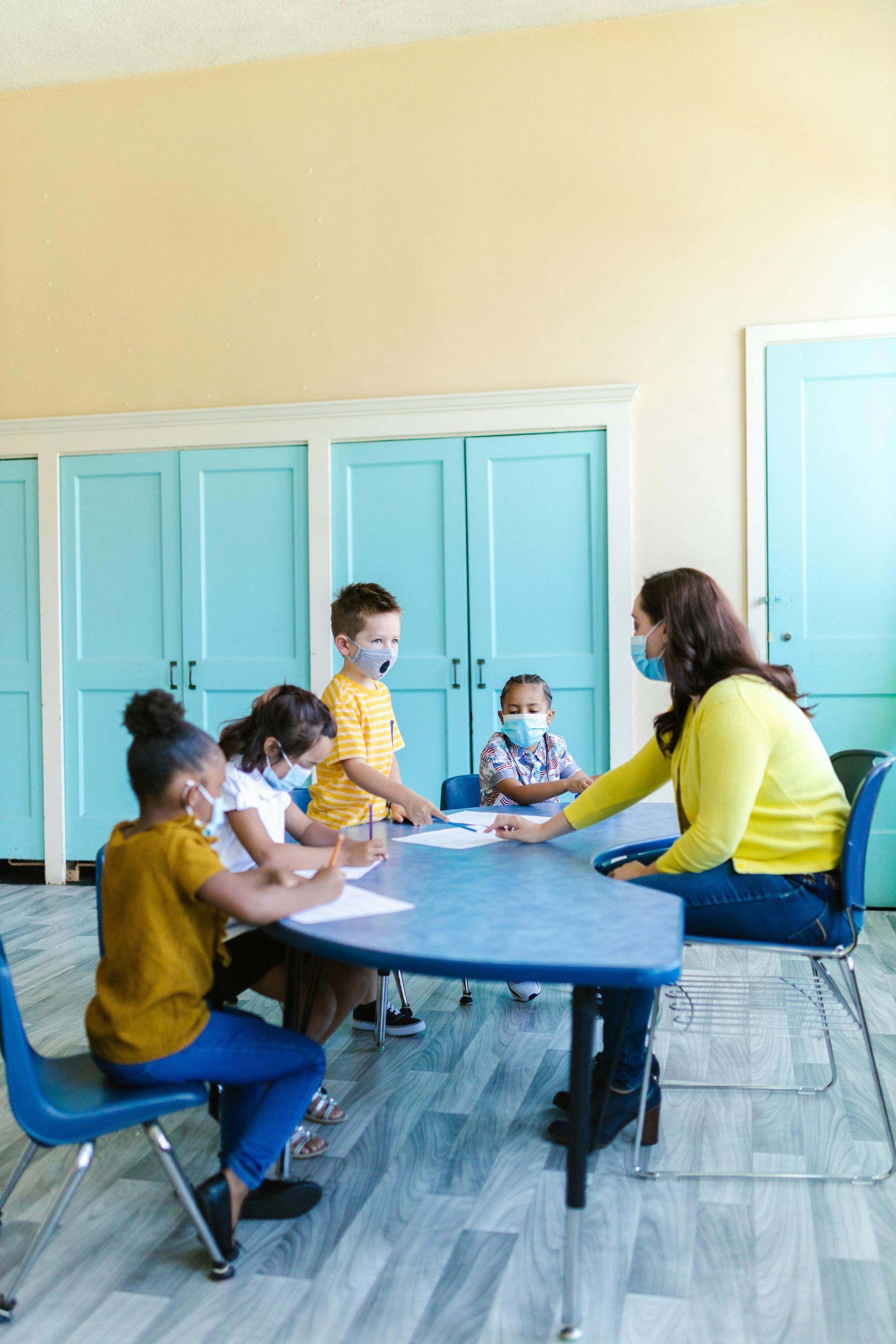 Children and teacher wearing masks at a blue table, writing. Classroom with blue doors, yellow wall.