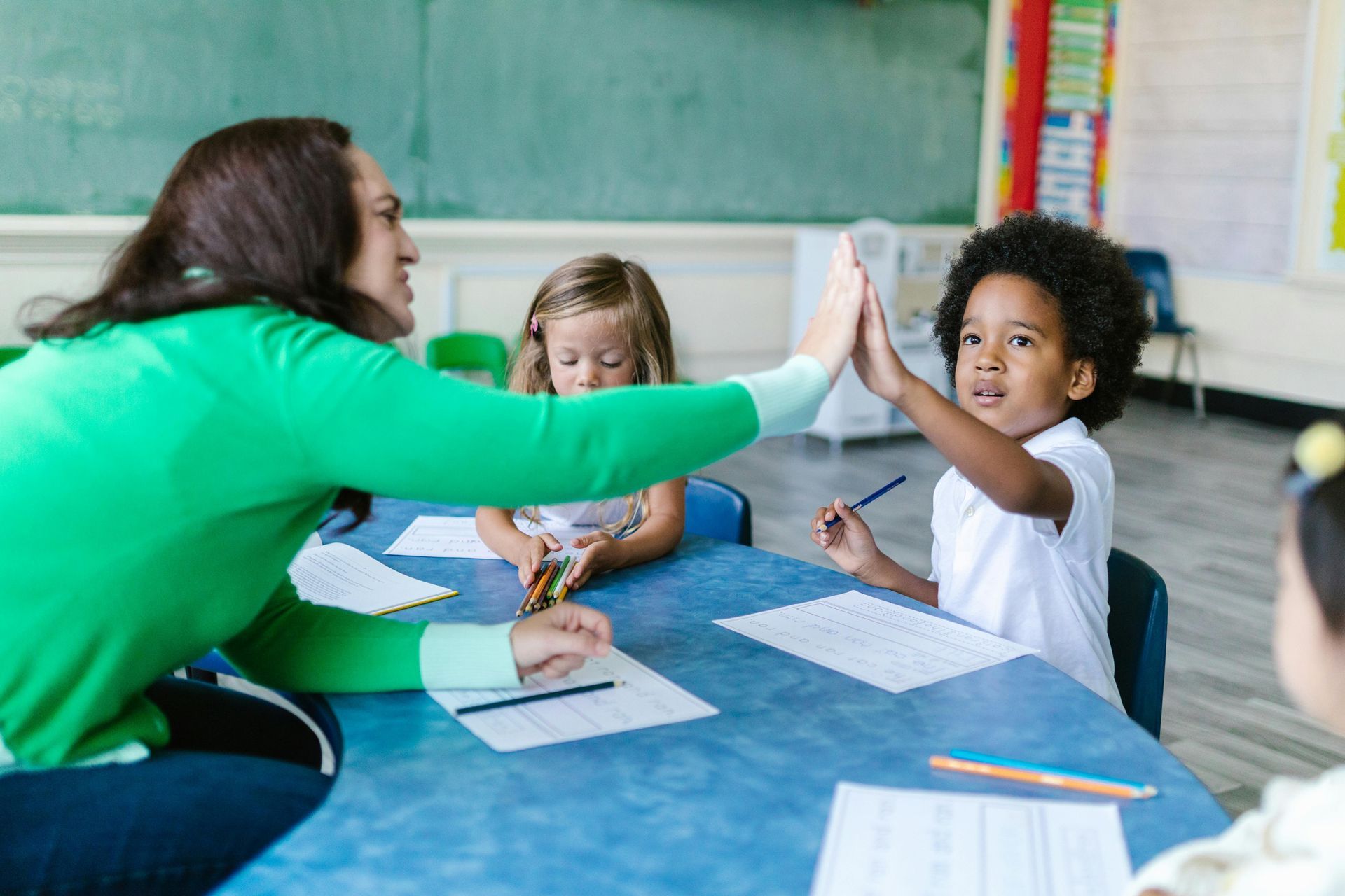 Teacher giving a high-five to a child at a blue table in a classroom; a girl is looking down.