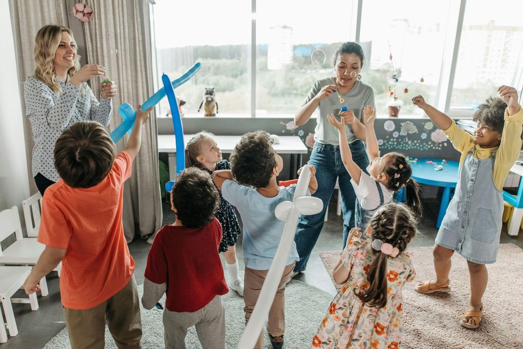 Children and teachers in a brightly lit playroom playing with balloons.