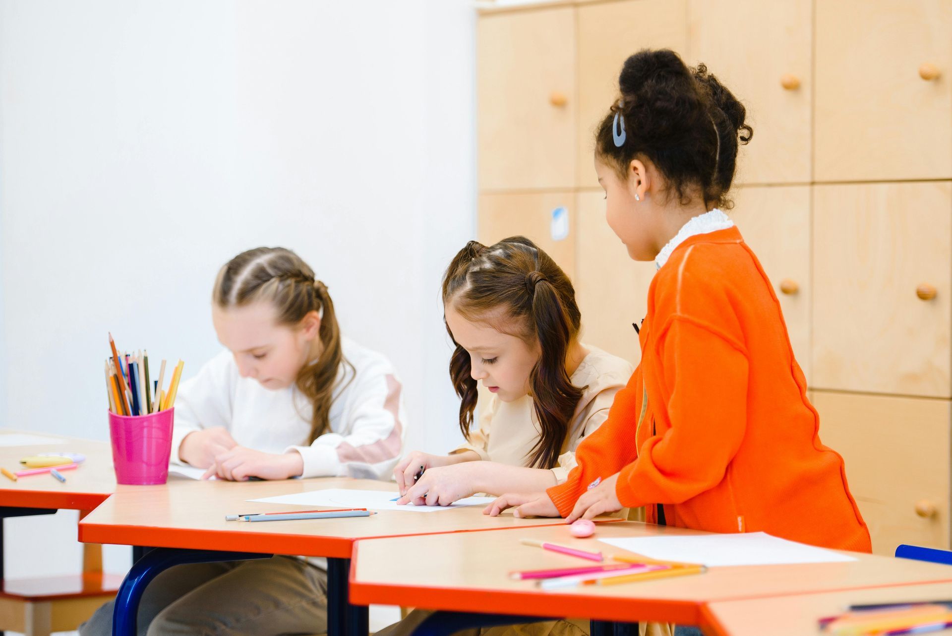 Three children seated at a desk, drawing with colored pencils in a classroom.