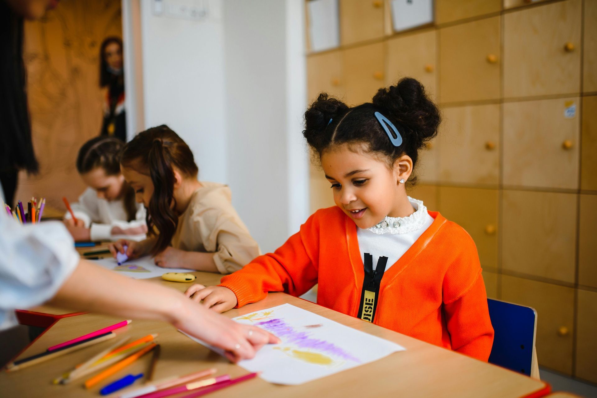 Girl in orange cardigan at a desk coloring with teacher's hand pointing at her drawing. Other children at desks in background.