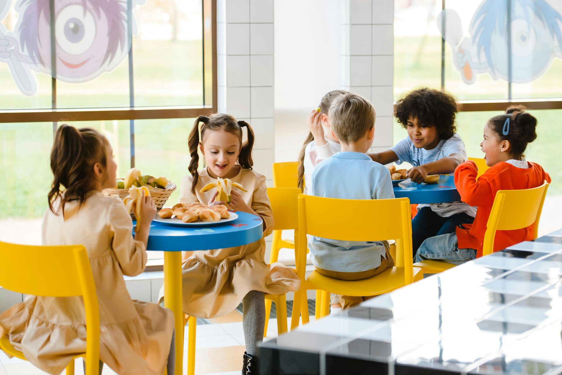 Children eating at blue tables in a brightly lit room with yellow chairs, some by windows with cartoon designs.