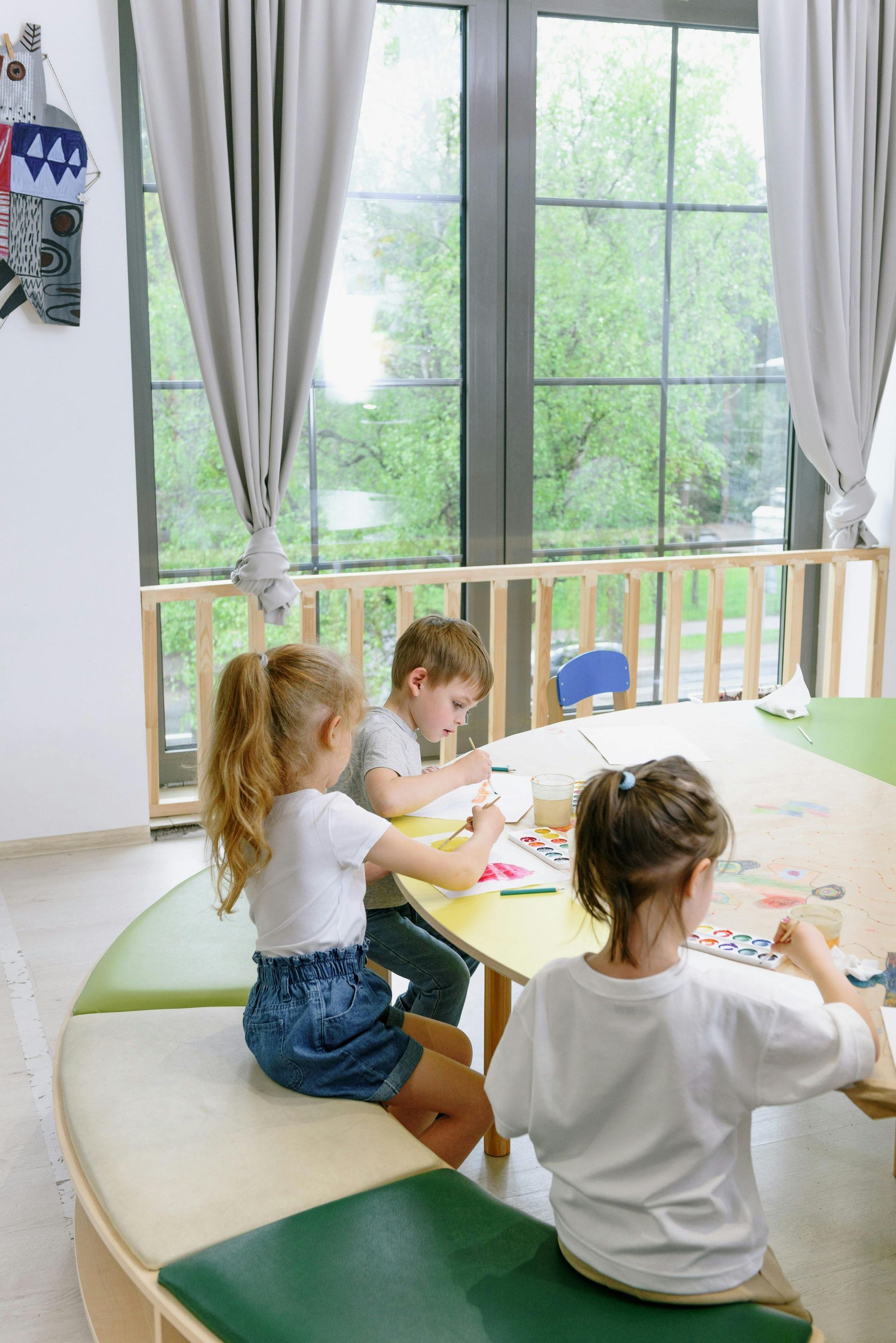 Three children seated at a table drawing inside a classroom, with a window and curtains in the background.