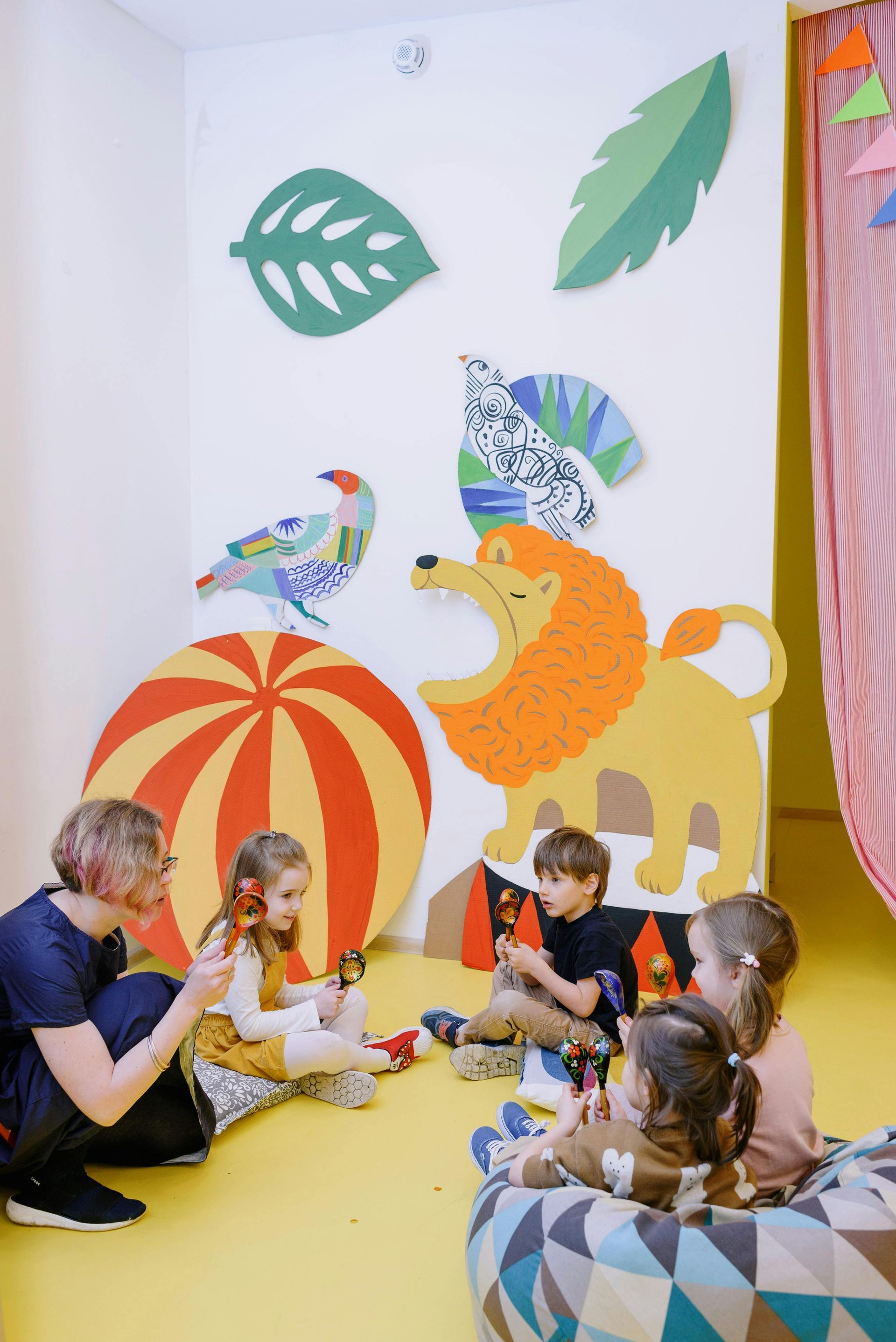 Children and adult in a colorful playroom with wall decor; playing with toys, sitting on pillows.