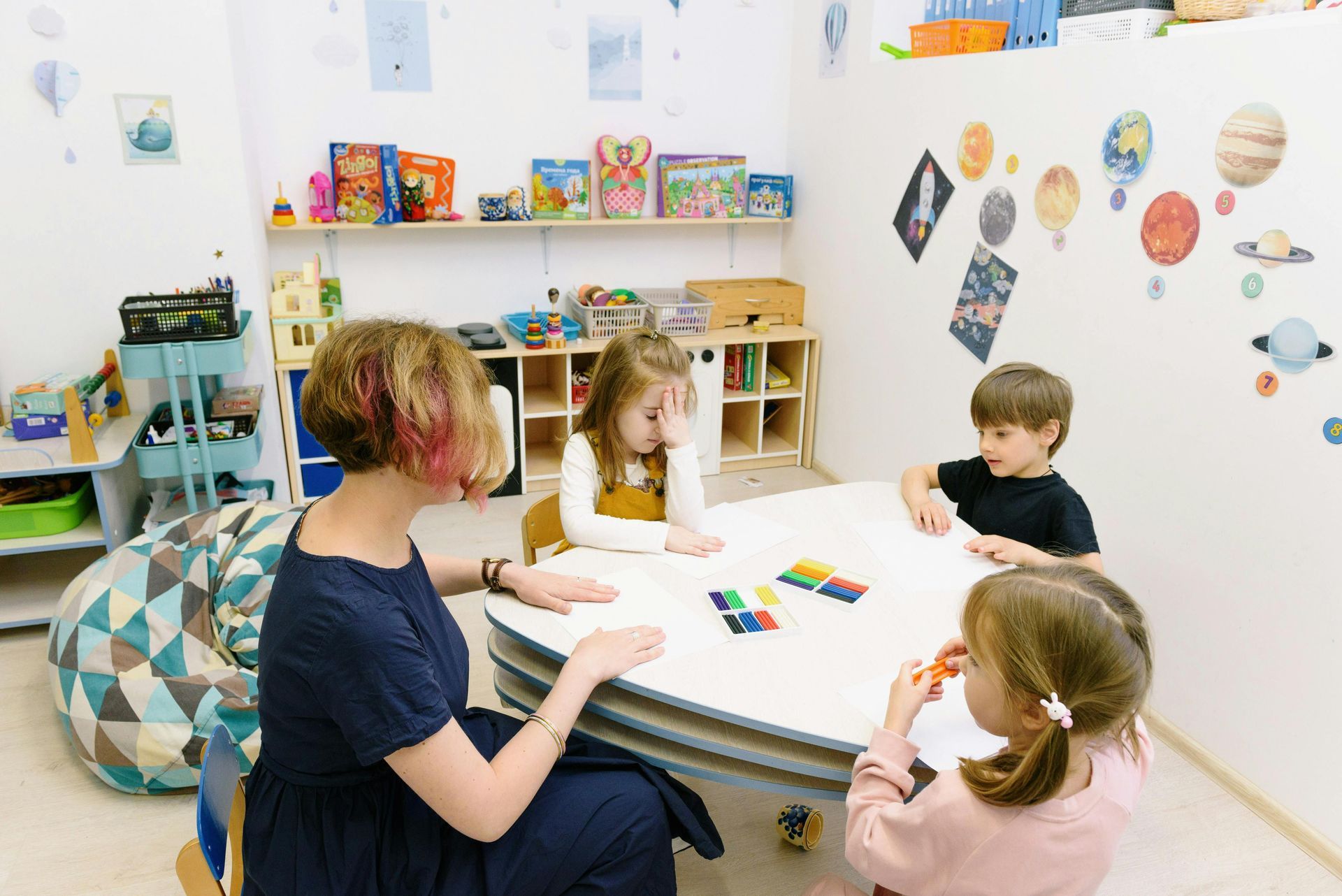 Woman and three children seated at a table, playing with colorful blocks in a playroom.