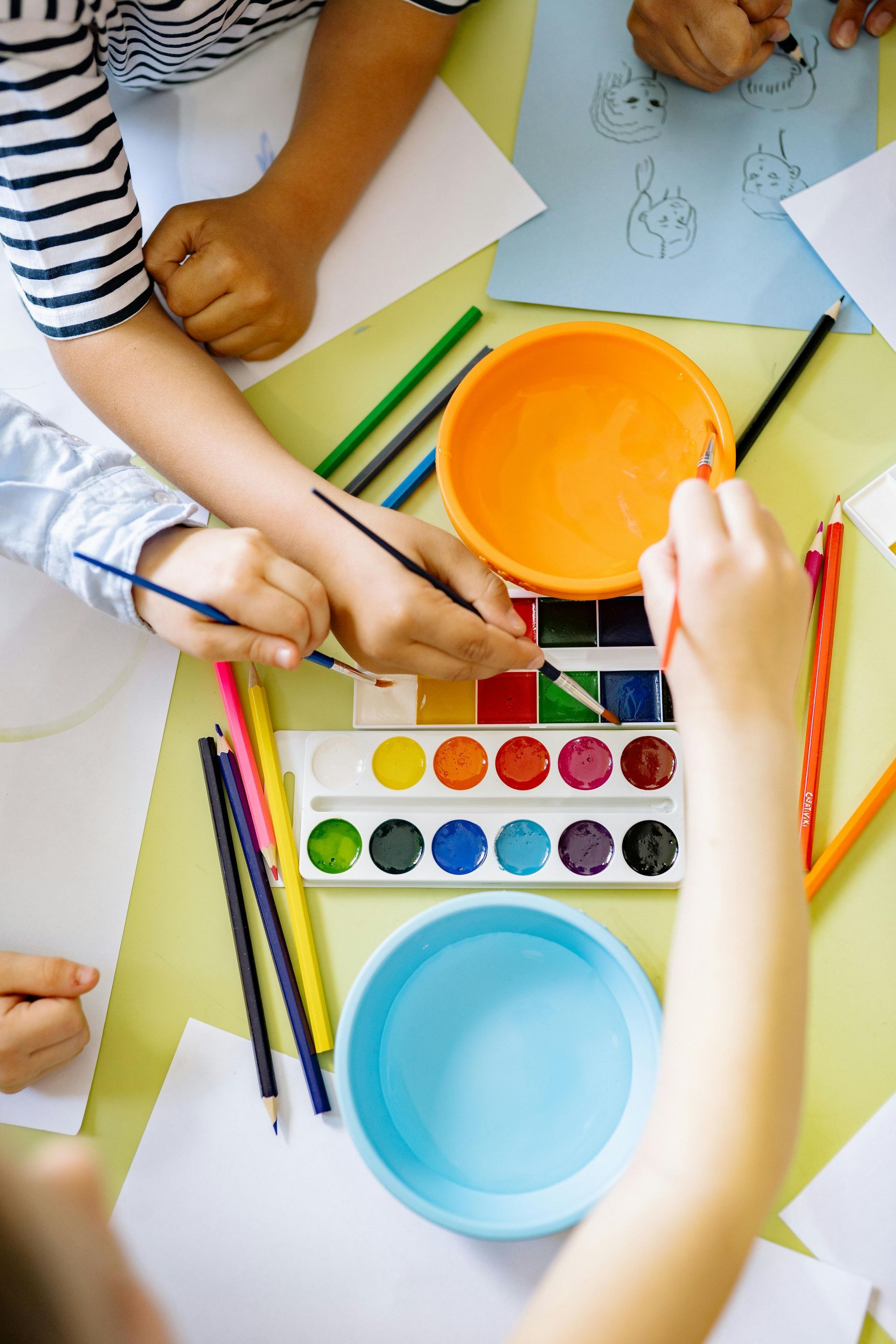 Children painting with watercolors at a table: hands using brushes, colors, and cups of water.