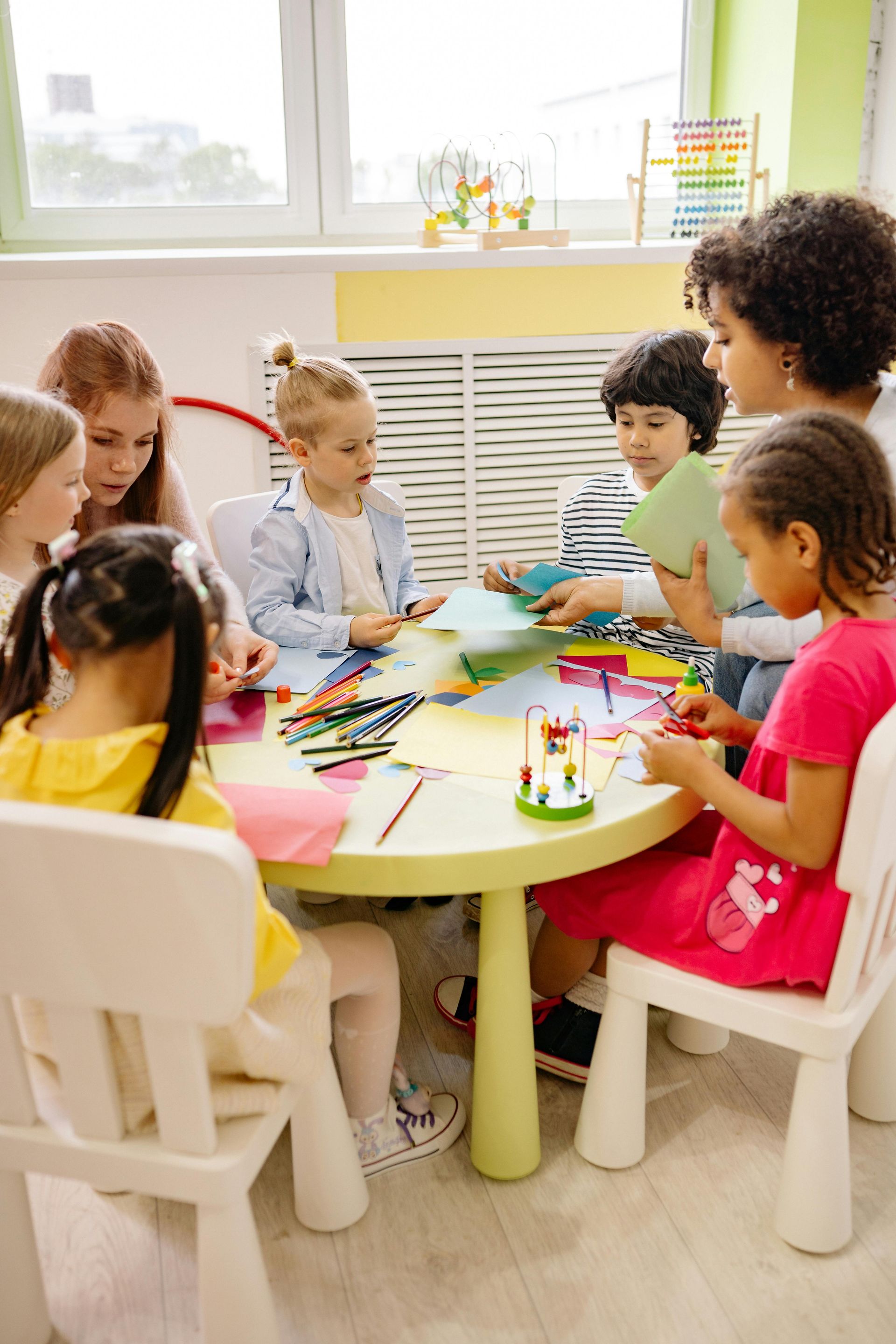 Children and a teacher at a table doing a craft project in a brightly lit classroom.