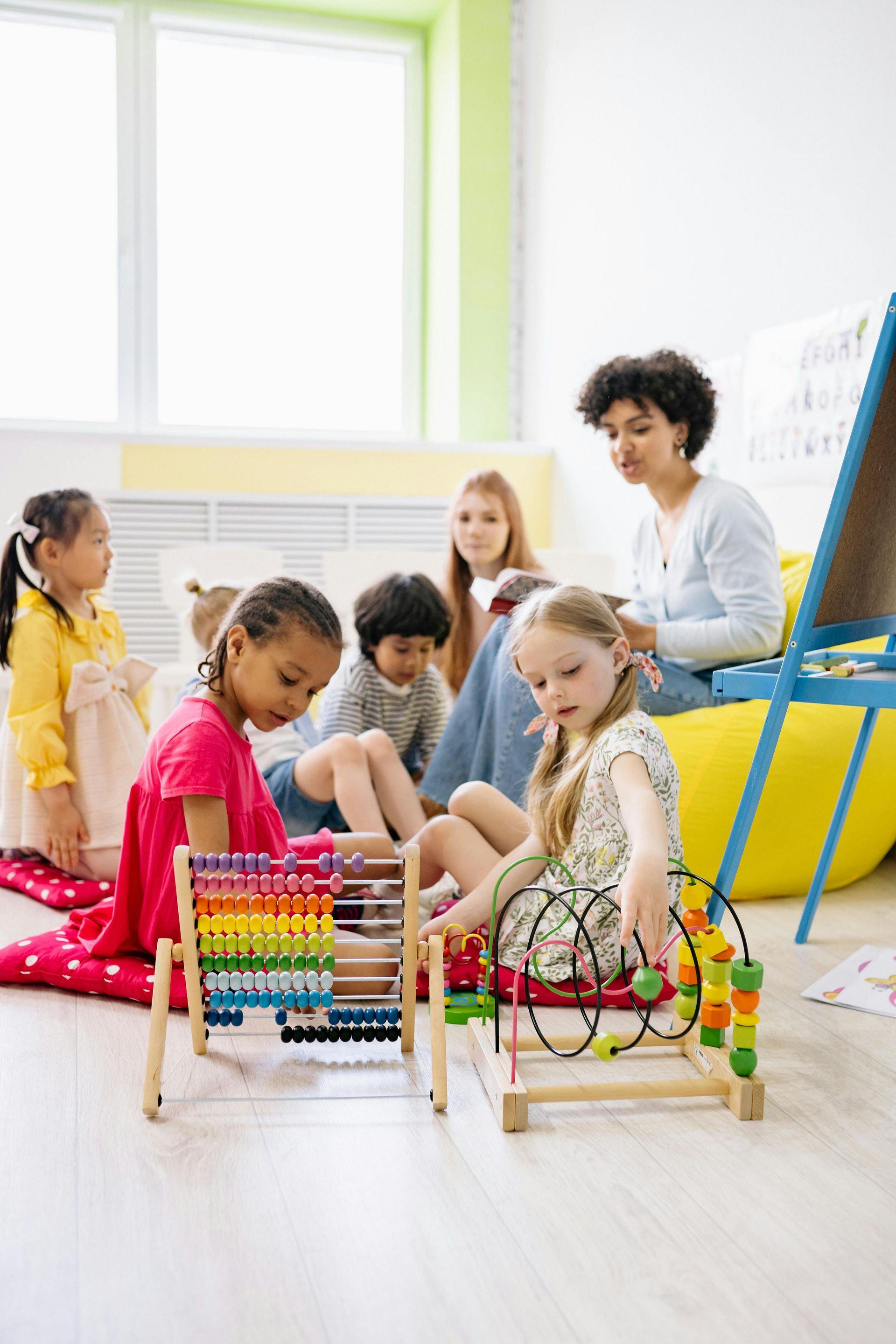 Children and teacher in a classroom with toys; one child uses an abacus, another uses a bead maze.
