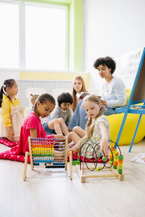 Children and teacher in a classroom with toys; one child uses an abacus, another uses a bead maze.