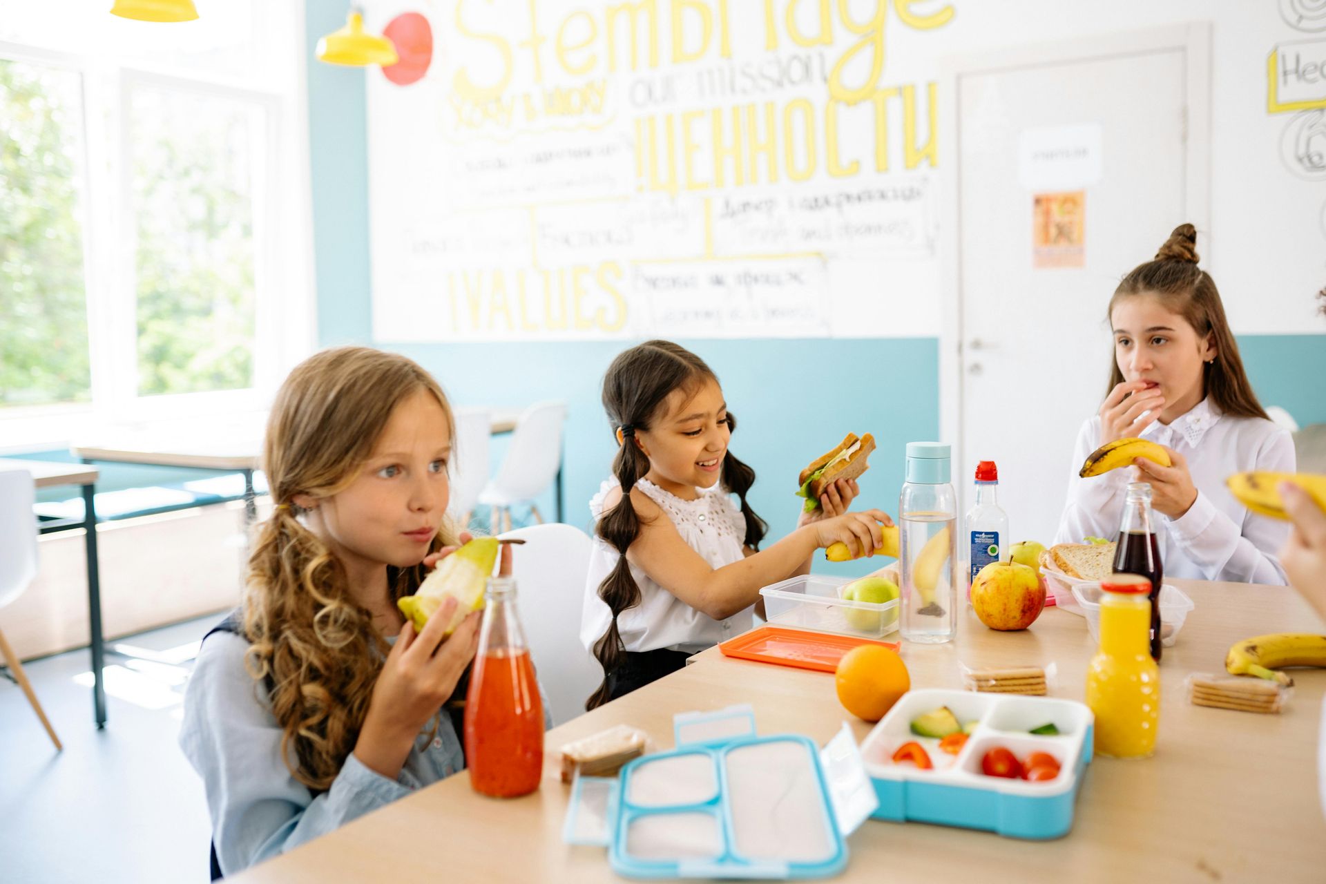 Children eating lunch at a table in a bright classroom. Includes sandwiches, fruit, and drinks.