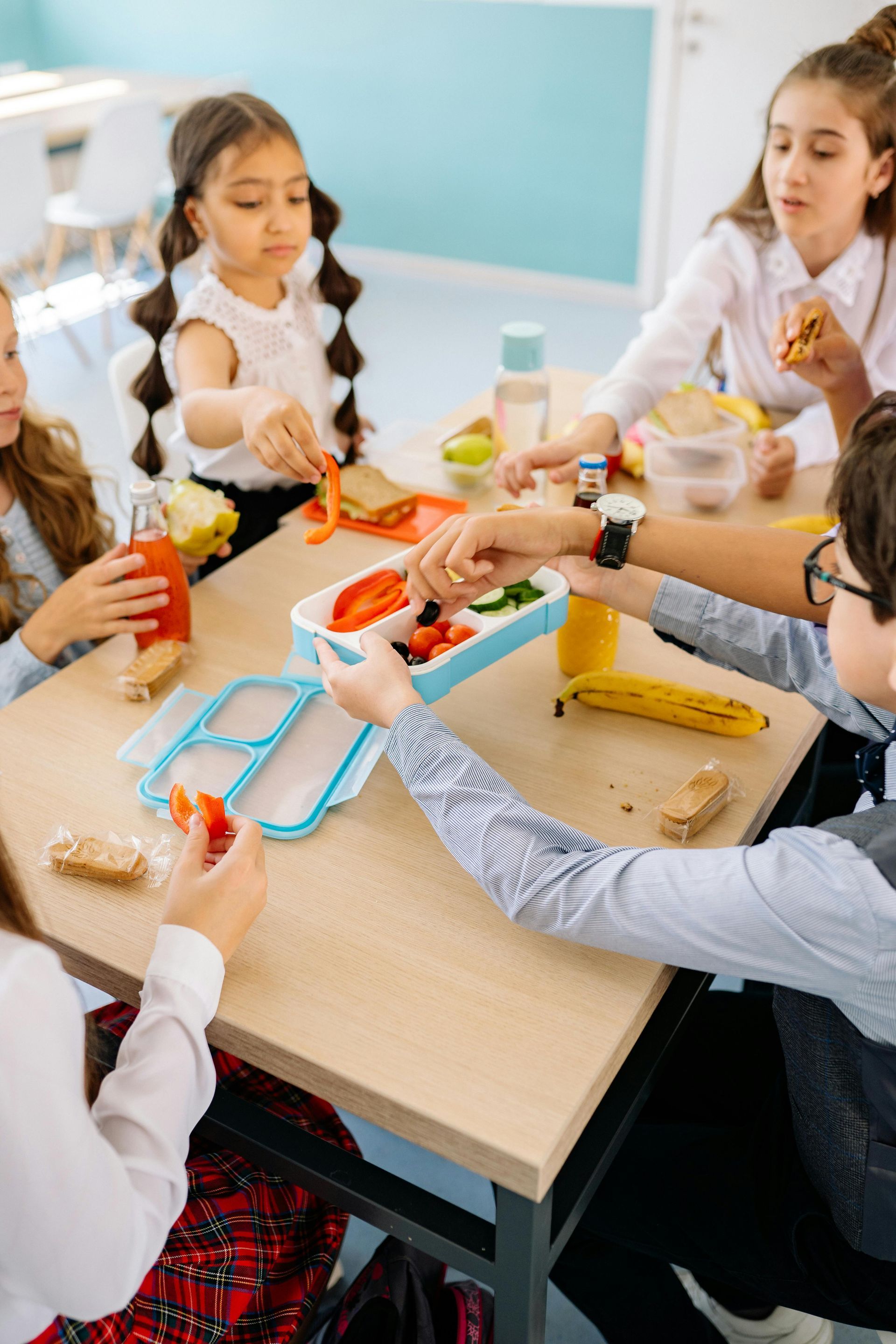 Children at a table eating lunch. They are reaching for items in a lunch box, holding food, and drinking juice.