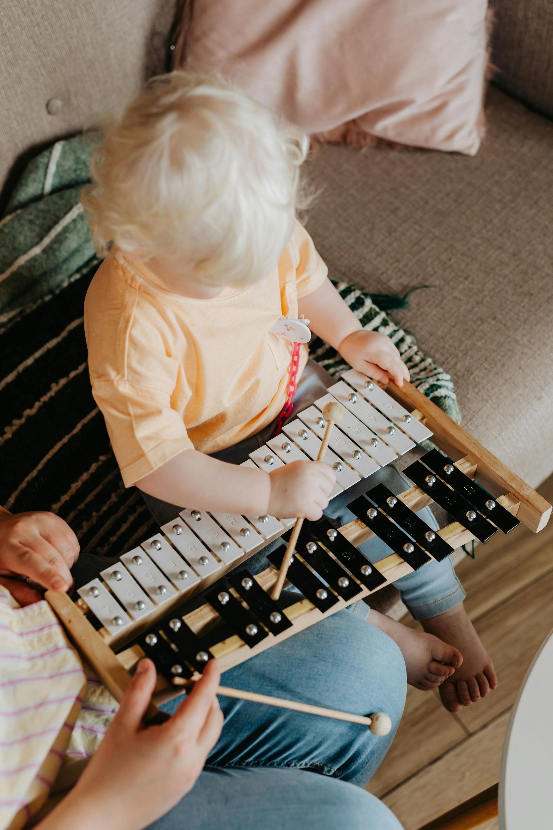 Child with blonde hair plays a xylophone on a person's lap; indoor setting.