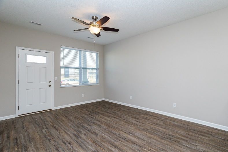 Empty living room with wood floors, light gray walls, and a ceiling fan. A door and window are on the left.