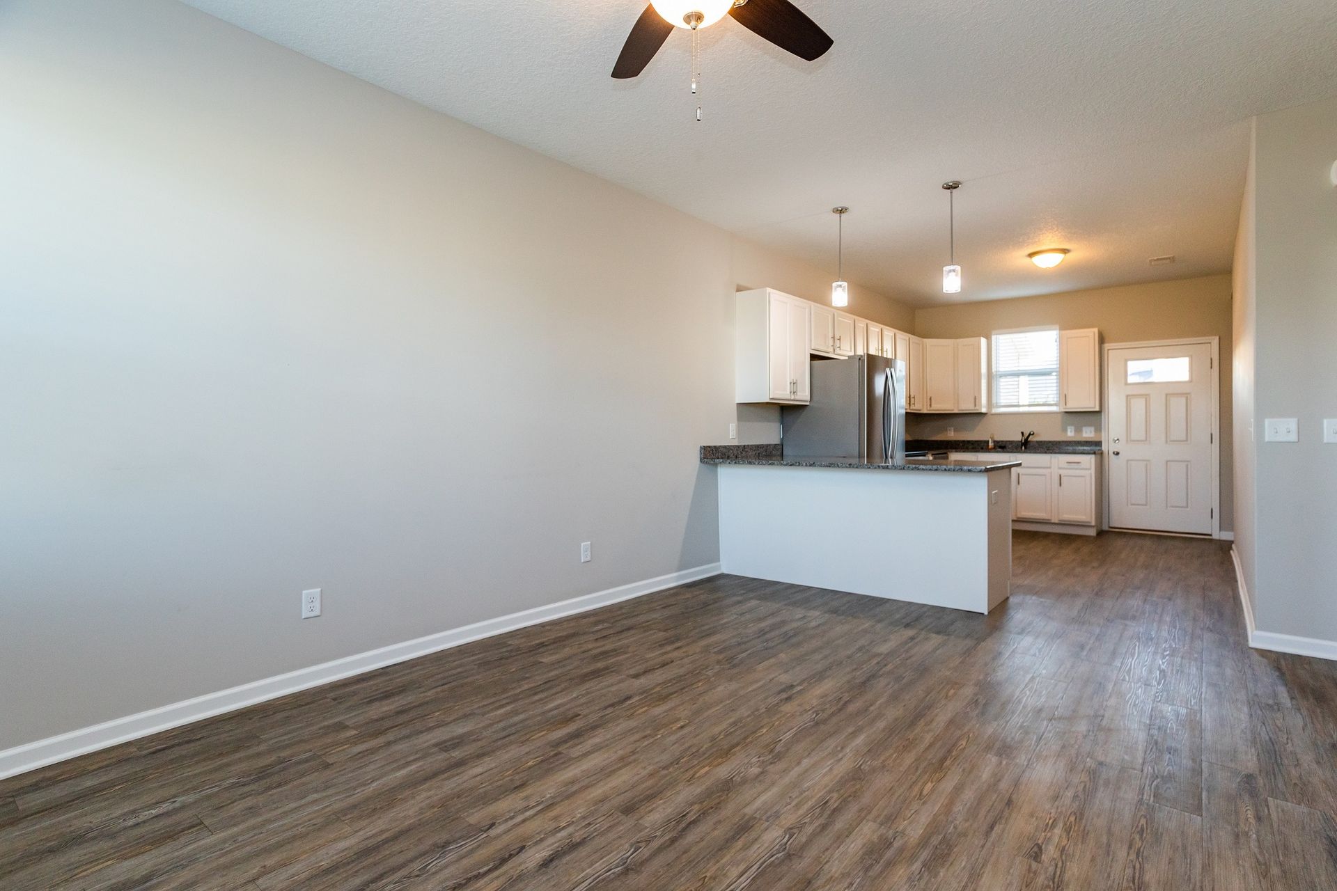 Empty living space with kitchen, hardwood floors, and beige walls.
