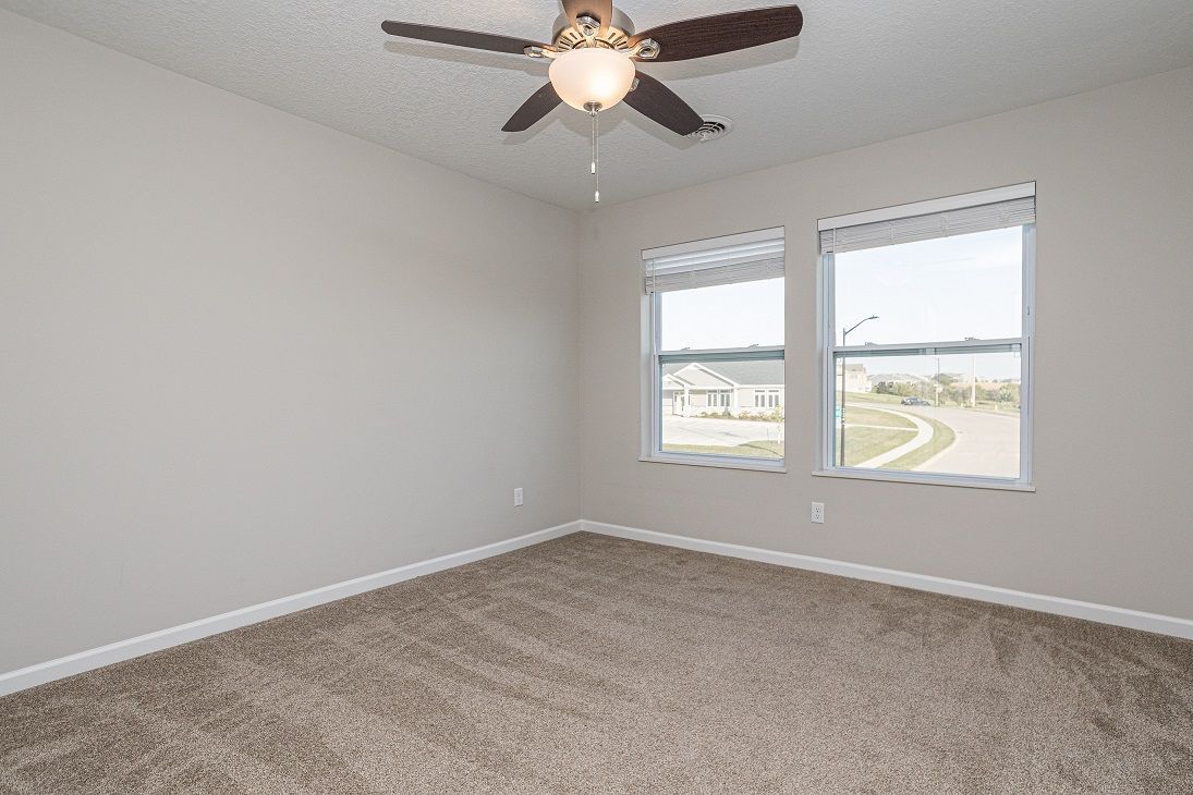 Empty bedroom with beige walls, brown carpet, two windows, and a ceiling fan.