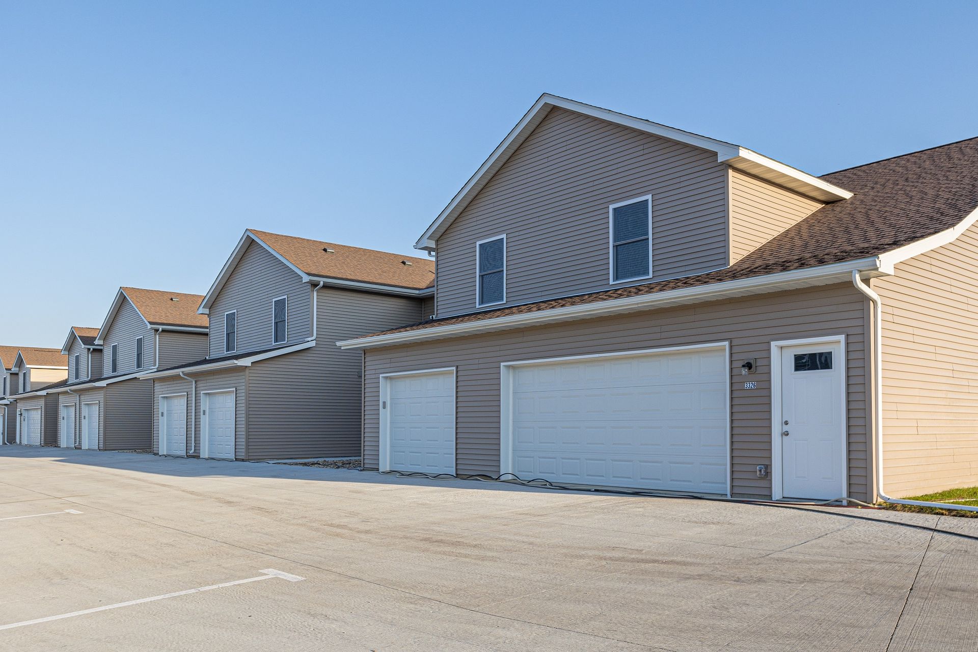 Row of tan apartment buildings with attached garages and asphalt parking lot under blue sky.