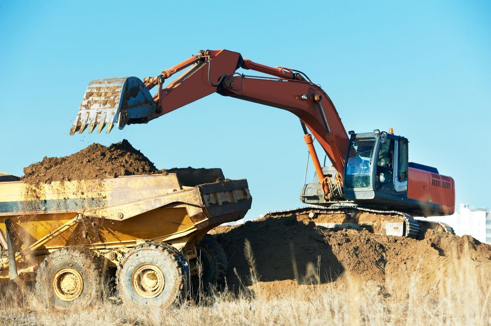 Excavator Loading Dirt Into a Yellow Dump Truck at a Construction Site Under a Clear Blue Sky — Mike Barlow Earthmoving Pty Ltd in Rockhampton, QLD