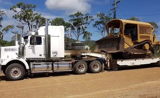 A White Semi Truck Is Carrying A Bulldozer Down A Dirt Road — Mike Barlow Earthmoving Pty Ltd in Rockhampton, QLD