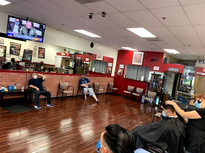 Inside a barbershop, people are getting haircuts and waiting, some wearing masks. Red walls and a TV are visible.