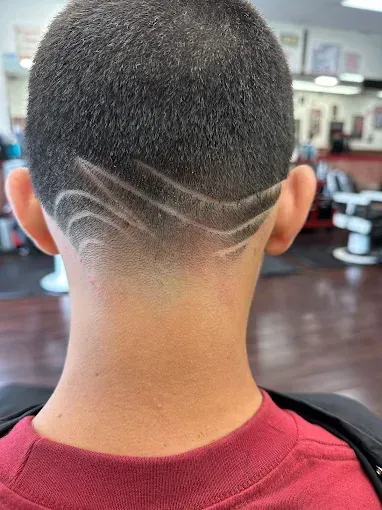 A person from the back, with a short haircut and etched design on the nape of the neck. The person wears a red shirt, in a barber shop.