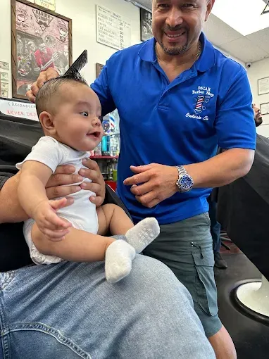 Barber giving a smiling baby a haircut at a shop. The barber wears a blue shirt, holding a comb near the baby’s head.