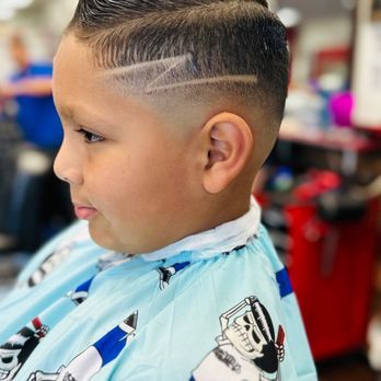 Young boy with a fade haircut, featuring a lightning bolt design. He's wearing a blue and white barber cape, and the background is a barbershop.