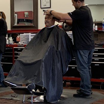 A man in a barber chair getting a haircut. A barber, wearing a black shirt and jeans, cuts the man's hair with clippers. The setting appears to be a barbershop.