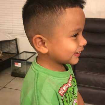Young boy with a fresh haircut, smiling, wearing a green shirt, and standing in front of a couch.