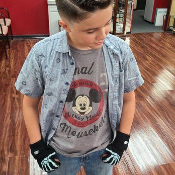 Boy with a side-parted haircut wearing a Mickey Mouse shirt, denim shirt, jeans, and fingerless skeleton gloves, standing indoors.