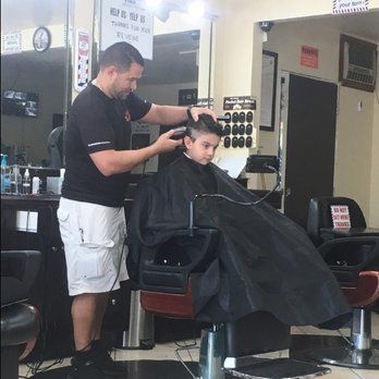 A barber giving a young boy a haircut in a barbershop. The barber is using clippers. The boy is wearing a black cape.