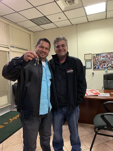 Two men smile indoors. One holds keys, seemingly having purchased a car. They stand near a desk in an office-like setting.