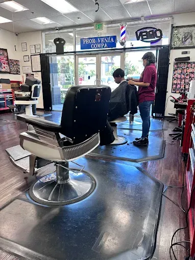 A barber cuts a customer's hair in a shop. The barber wears a red shirt. The shop has a retro barber chair.