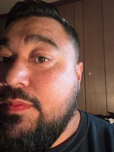 Close-up of a man with a dark beard and hair, looking at the camera. He is indoors, with brown skin and a neutral expression.